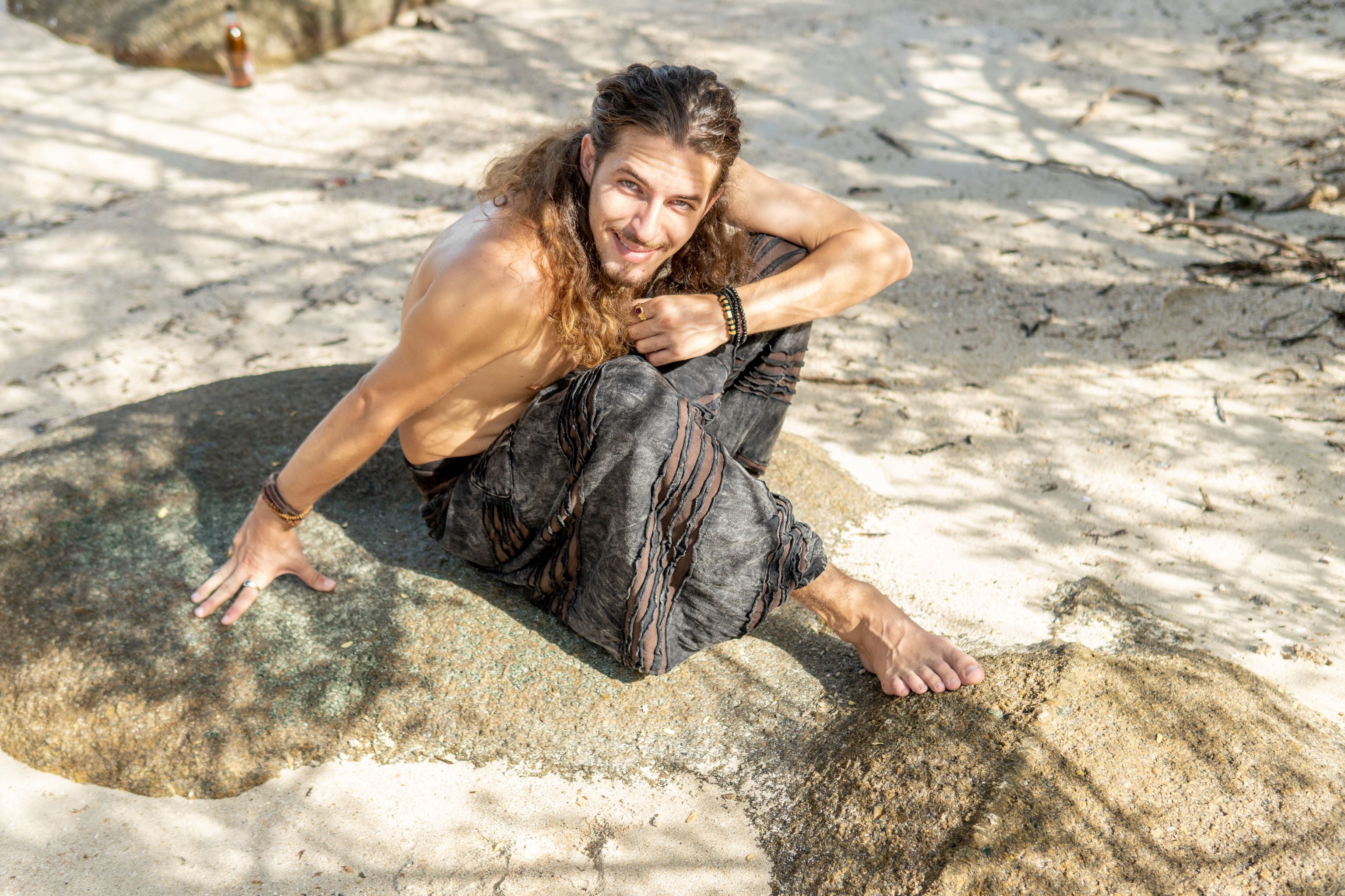 Man relaxing on a beach wearing charcoal grey stonewashed hippie trousers with a brown razorcut underlay, elasticated ankles and a relaxed drop crotch fit.
