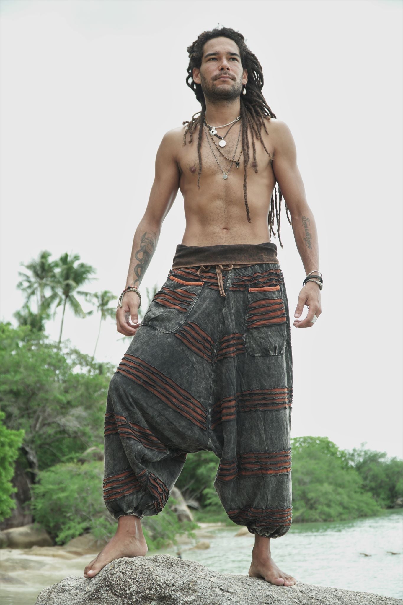 Man modelling charcoal stonewashed bohemian razorcut trousers with a rust orange underlayer on tropical beach rocks.