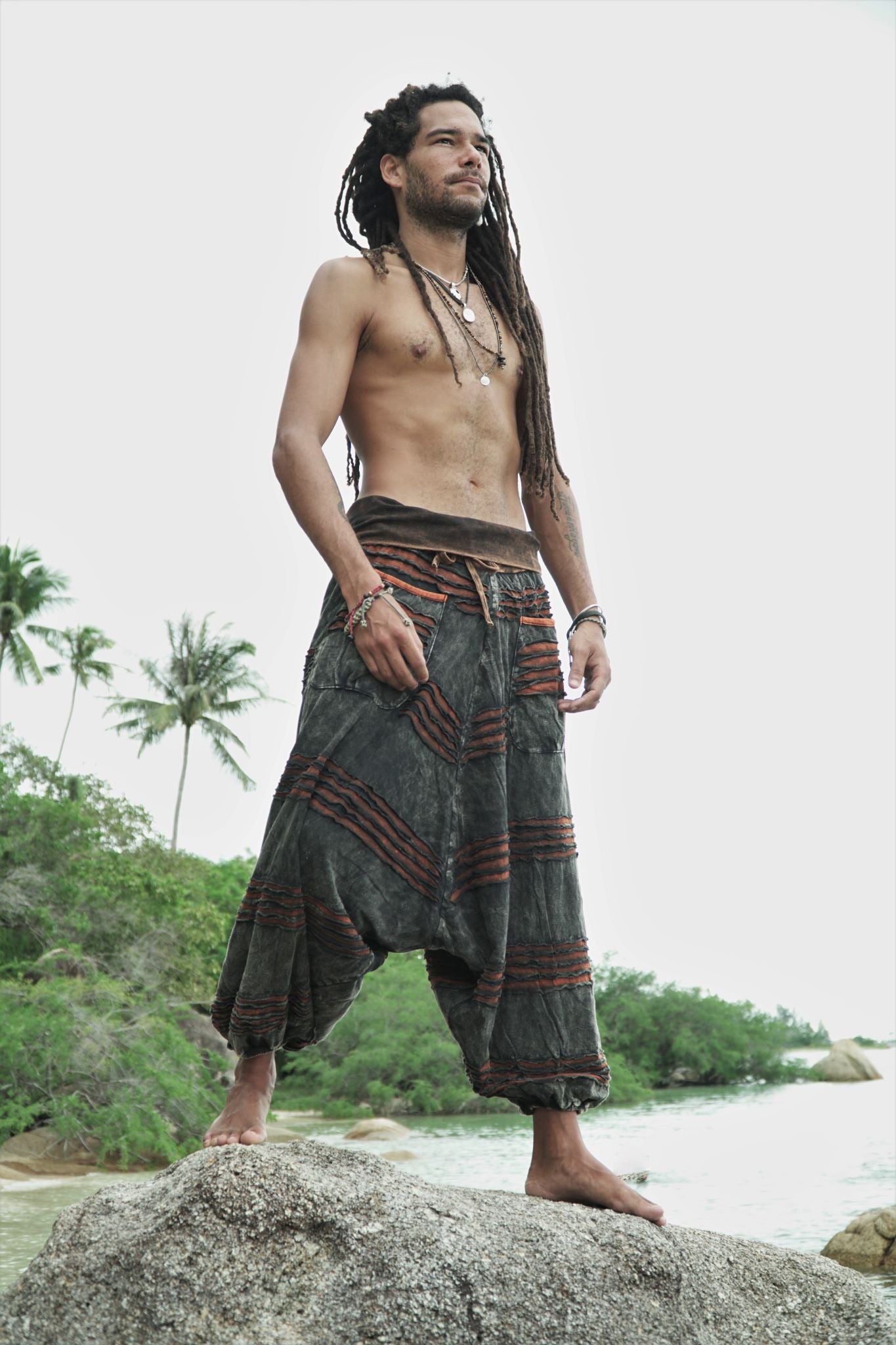 Man wearing charcoal stonewashed festival razorcut trousers with a rust orange underlayer on a tropical shoreline.