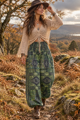 A woman walking on an outdoor mountain path wearing a pair of fuzzy green thick wide-leg trousers, a brown wide-brim hat, and brown boots.