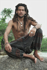 Man sitting on beach rocks wearing charcoal stonewashed hippie razorcut harem pants with a rust orange underlayer.