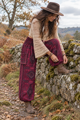 Woman wearing warm maroon patterned harem trousers with a shirred waistband and side pockets, adjusting her boot beside a dry-stone wall.