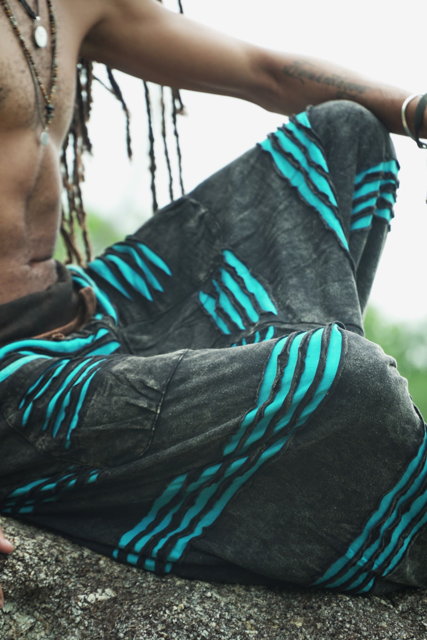 Close-up of charcoal stonewashed boho harem pants showing the handcrafted razorcut design with vivid blue underlayer detail and textured cotton fabric, worn cross-legged on a rock.