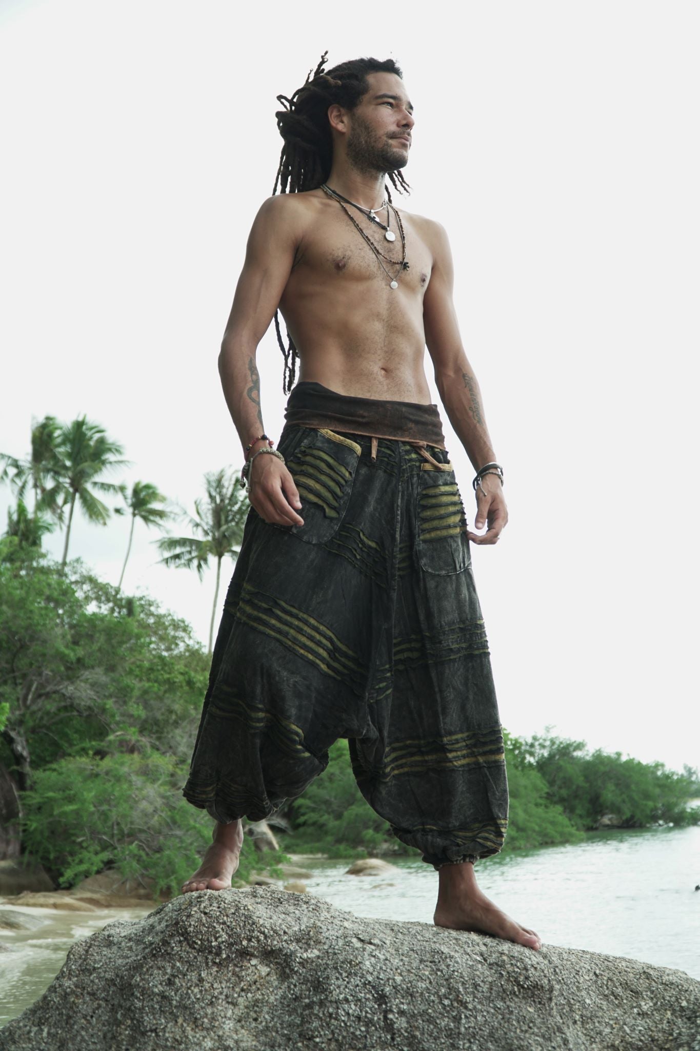 Male model with dreadlocks standing on a beach rock wearing charcoal stonewashed boho harem pants with a green razorcut underlayer, showcasing the wide leg drape and drop crotch silhouette against a tropical backdrop.