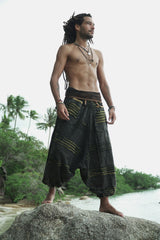 Male model with dreadlocks standing on a beach rock wearing charcoal stonewashed boho harem pants with a green razorcut underlayer, showcasing the wide leg drape and drop crotch silhouette against a tropical backdrop.