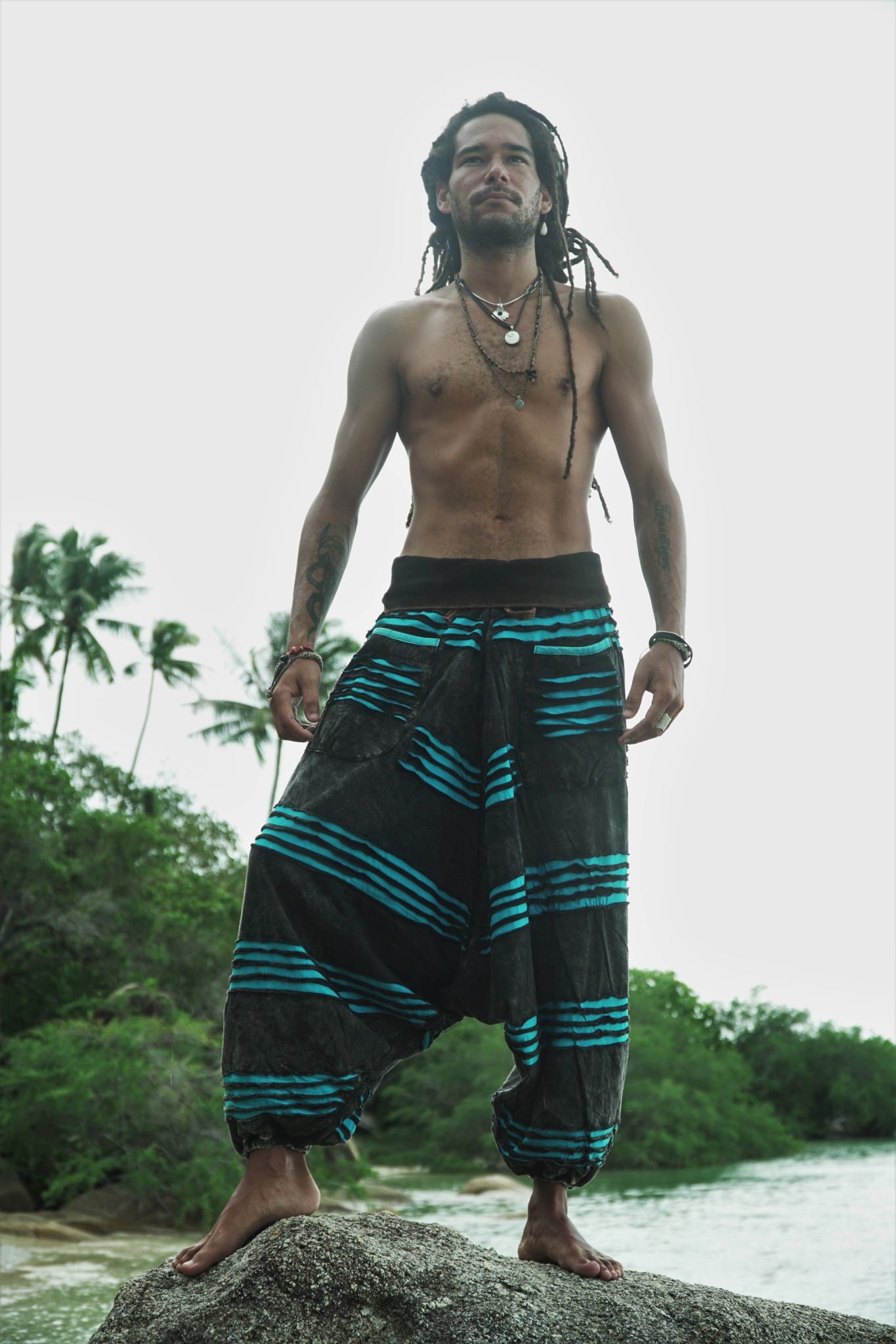 Full-length view of a male model with dreadlocks standing on a beach rock wearing charcoal stonewashed boho trousers with a vivid blue razorcut underlayer, set against a tropical palm tree backdrop.