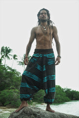 Full-length view of a male model with dreadlocks standing on a beach rock wearing charcoal stonewashed boho trousers with a vivid blue razorcut underlayer, set against a tropical palm tree backdrop.