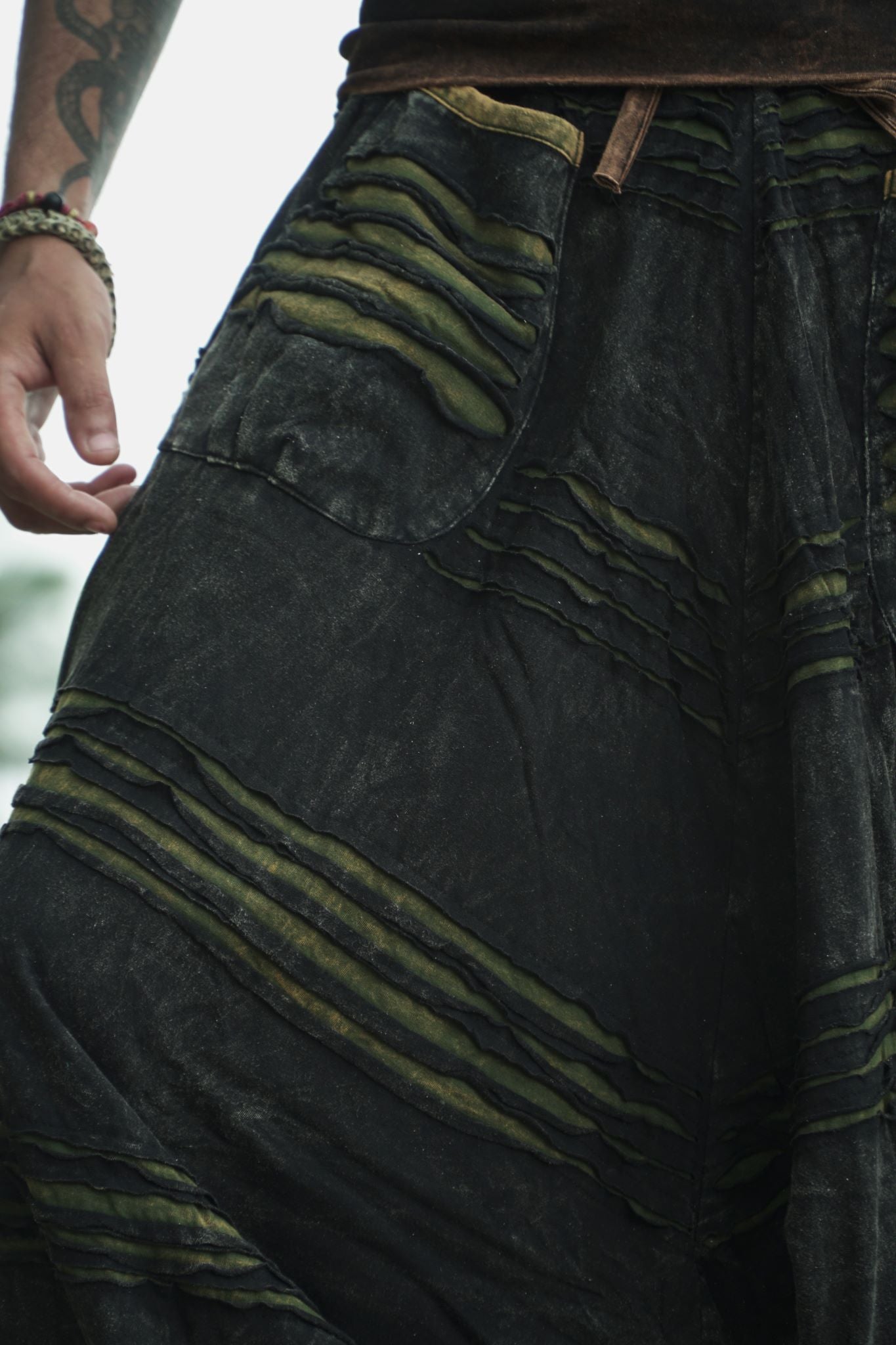 Close-up of men's charcoal grey stonewashed boho trousers showing razorcut pocket detail with a green underlayer and stonewashed fabric on a tattooed male model.