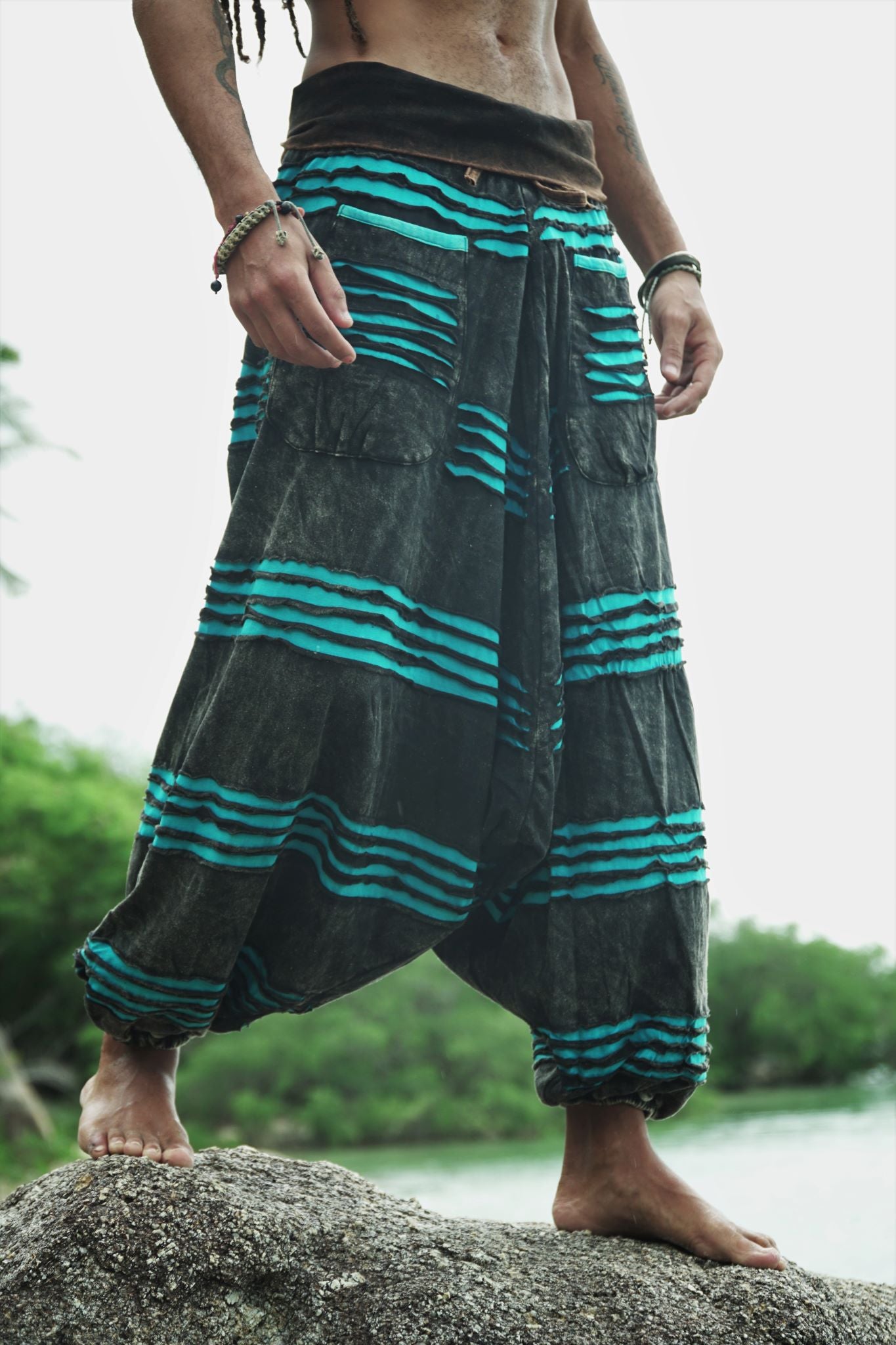 Waist-to-feet hero shot of men's charcoal stonewashed drop crotch  harem trousers with a vivid blue razorcut underlayer, showing oversized pockets, wide leg drape and elasticated ankles on a beach rock.