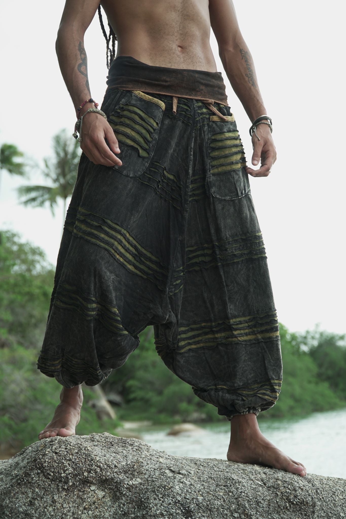 Waist-to-feet view of men's charcoal stonewashed drop crotch trousers with a green razorcut underlayer, showing oversized pockets, elasticated ankles and relaxed fit on a tropical beach rock.