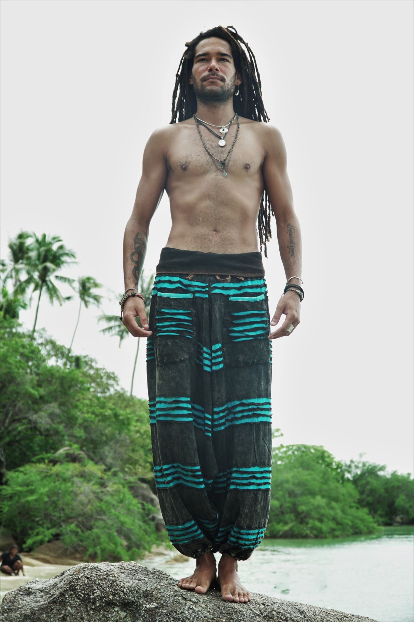 Male model with dreadlocks standing barefoot on a beach rock wearing charcoal stonewashed festival trousers with a bright blue razorcut underlayer, with a tropical river and palm trees in the background.