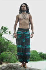 Male model with dreadlocks standing barefoot on a beach rock wearing charcoal stonewashed festival trousers with a bright blue razorcut underlayer, with a tropical river and palm trees in the background.