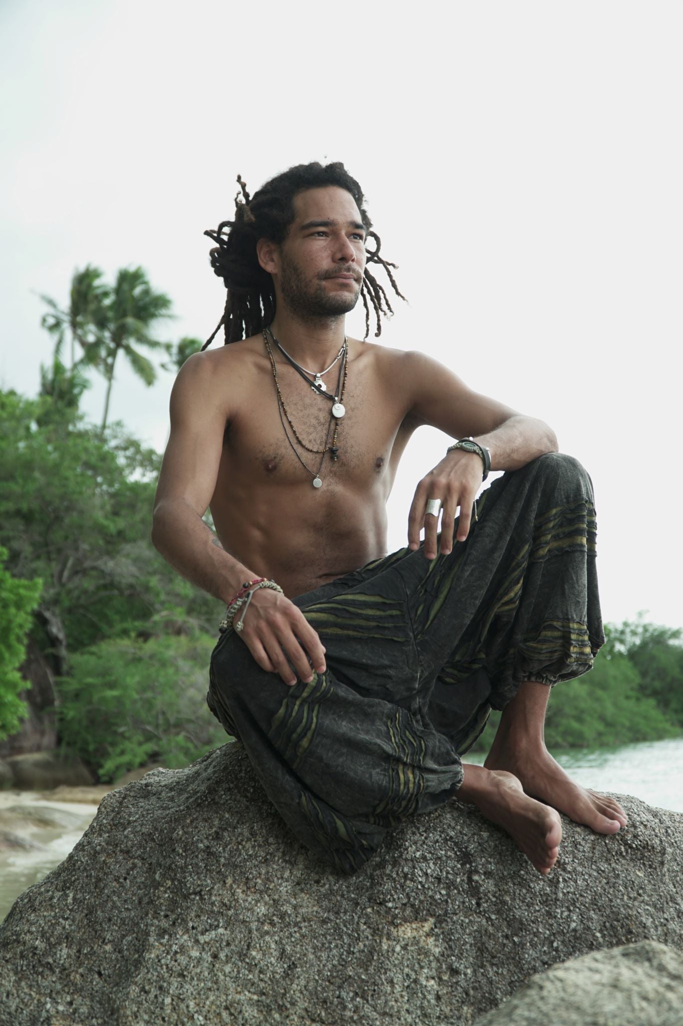 Male model with dreadlocks sitting cross-legged on a beach rock wearing charcoal stonewashed festival trousers with a green razorcut underlayer, shown in a relaxed bohemian lifestyle pose with a tropical backdrop.