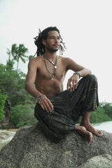 Male model with dreadlocks sitting cross-legged on a beach rock wearing charcoal stonewashed festival trousers with a green razorcut underlayer, shown in a relaxed bohemian lifestyle pose with a tropical backdrop.