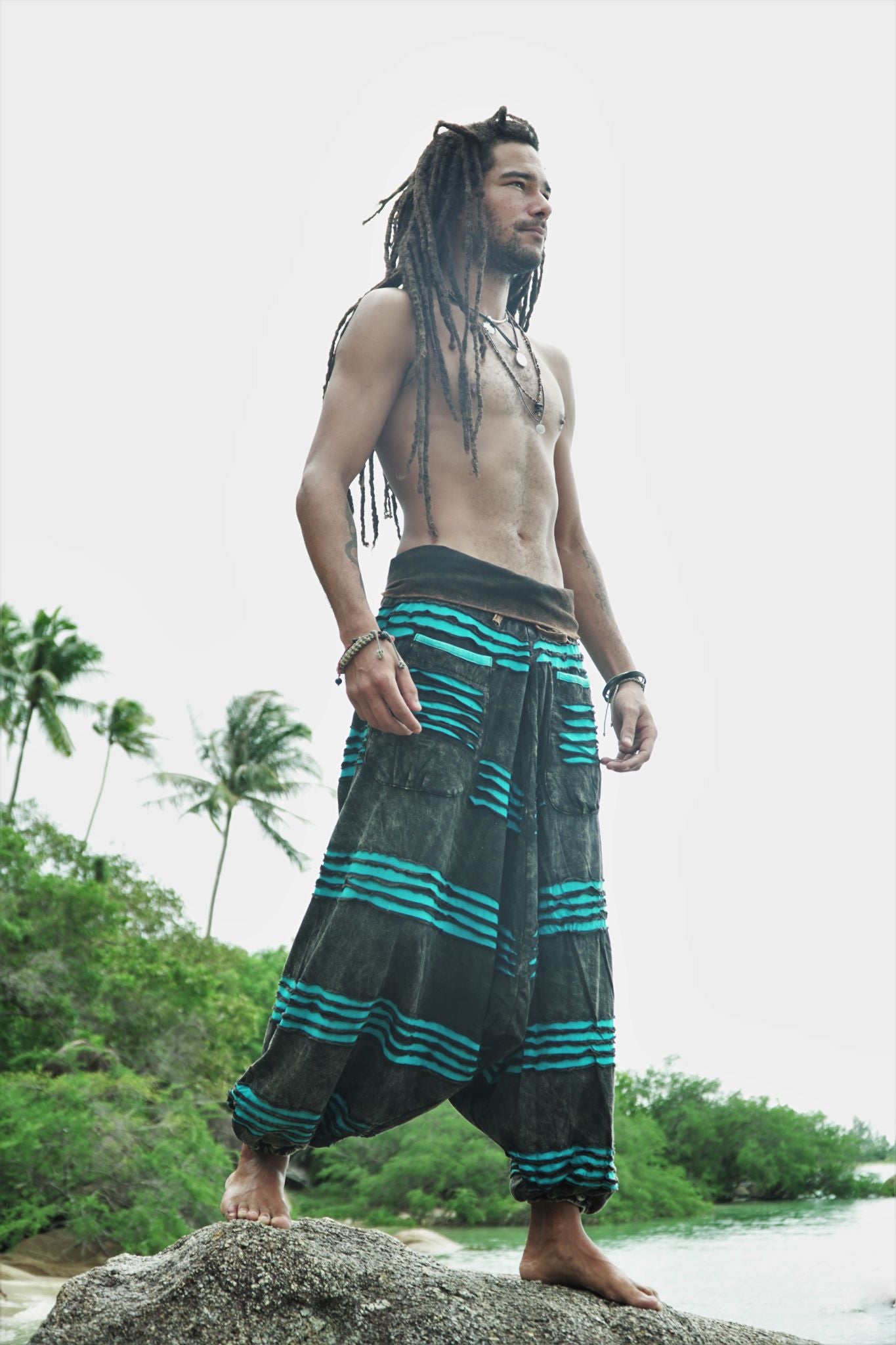 Side view of a male model with dreadlocks standing on a beach rock wearing charcoal stonewashed genie pants with a vivid blue razorcut underlayer, with palm trees and a tropical river in the background.
