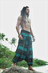 Side view of a male model with dreadlocks standing on a beach rock wearing charcoal stonewashed genie pants with a vivid blue razorcut underlayer, with palm trees and a tropical river in the background.