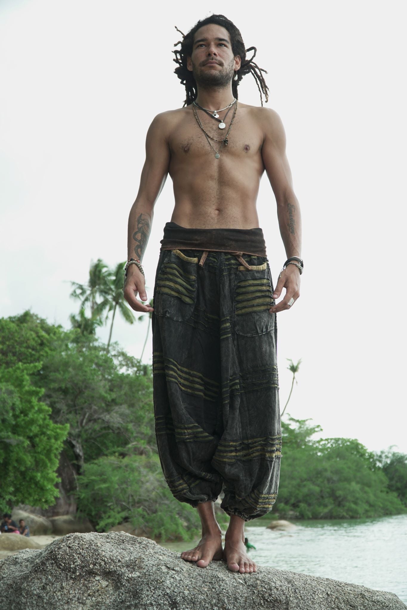Male model with dreadlocks standing on beach rocks wearing charcoal stonewashed genie pants with a green razorcut underlayer, showing the relaxed drop crotch fit and elasticated ankles in a tropical beach setting.