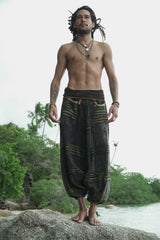 Male model with dreadlocks standing on beach rocks wearing charcoal stonewashed genie pants with a green razorcut underlayer, showing the relaxed drop crotch fit and elasticated ankles in a tropical beach setting.