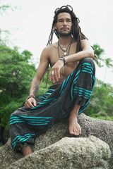 Male model with dreadlocks sitting on rocks wearing grey stonewashed razorcut harem pants with a vivid blue underlayer, in a relaxed bohemian pose with a tropical backdrop.