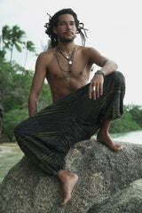 Male model sitting on a rock wearing charcoal stonewashed razorcut harem pants with a green underlayer that flashes through each cut, shown in a relaxed front view on a tropical beach.