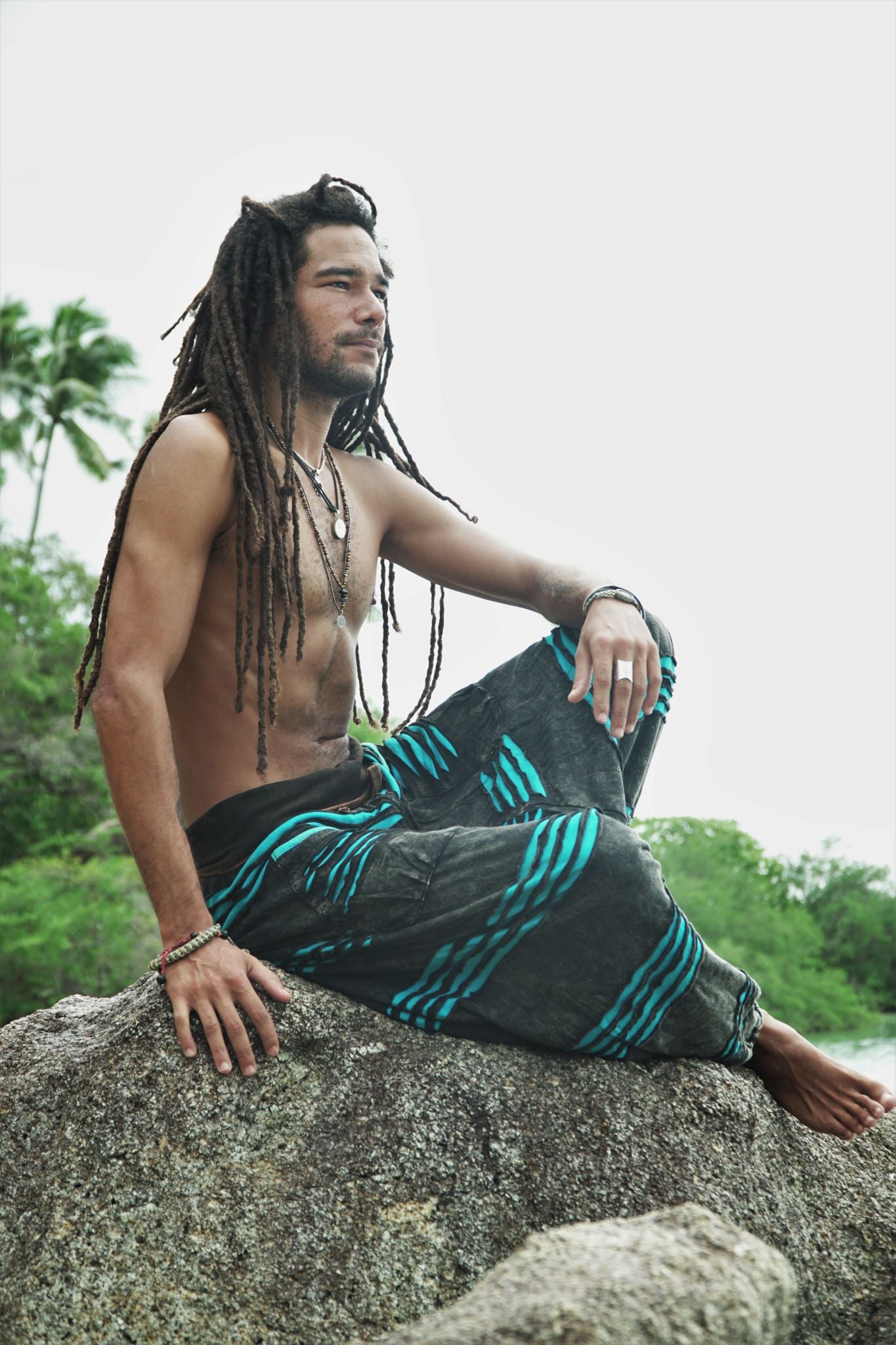 Male model with dreadlocks sitting on a rock in profile, wearing grey stonewashed hippie pants with a bright blue razorcut underlayer, in a relaxed bohemian pose with a tropical beach backdrop.