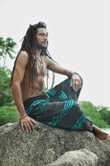 Male model with dreadlocks sitting on a rock in profile, wearing grey stonewashed hippie pants with a bright blue razorcut underlayer, in a relaxed bohemian pose with a tropical beach backdrop.