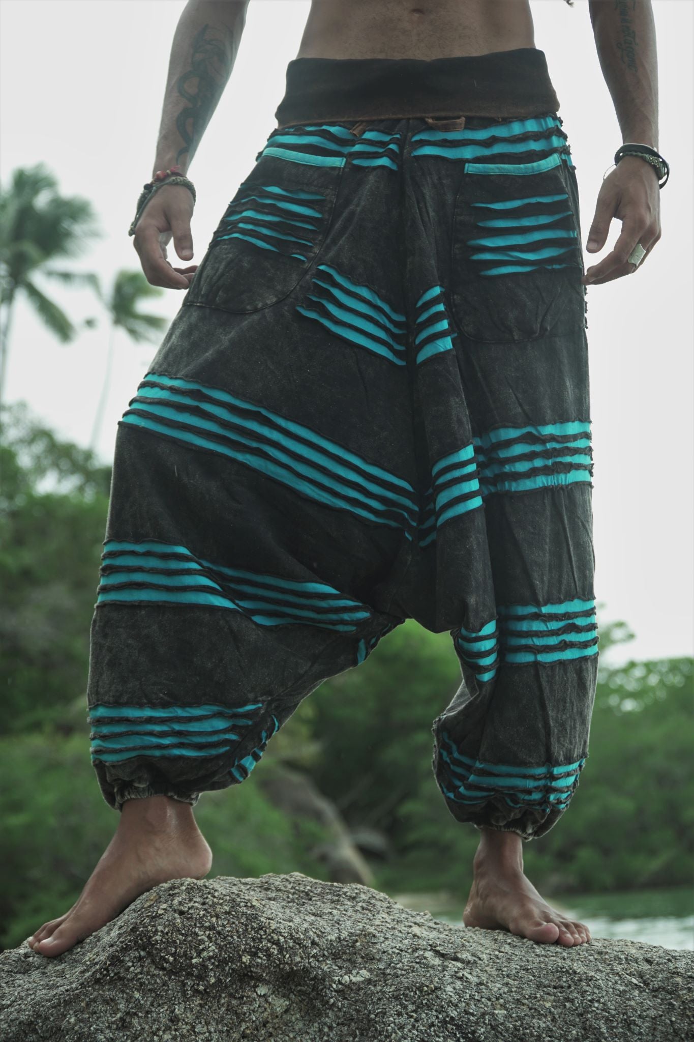 Waist-to-feet front view of men's charcoal grey stonewashed hippie trousers with a bright blue razorcut underlayer, showing the drop crotch drape, oversized pockets and elasticated ankles on a beach rock.