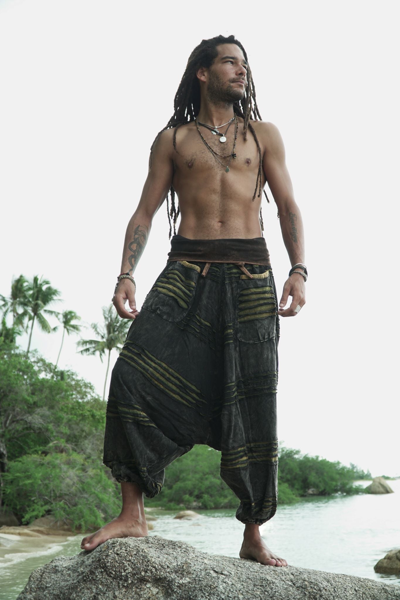 Male model with dreadlocks standing on beach rocks wearing charcoal stonewashed hippie trousers with a green razorcut underlayer, showing the full drop crotch silhouette in a tropical setting.