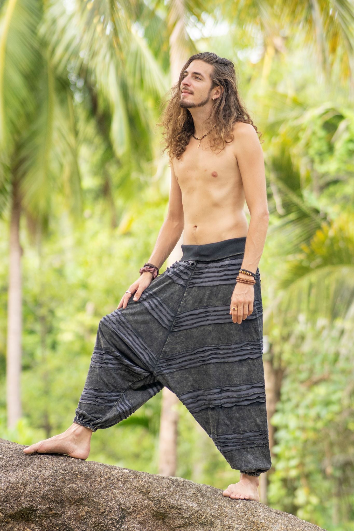 Man wearing grey stonewashed cotton razorcut genie pants with a relaxed drop crotch fit, layered razorcut panels, elasticated waistband and ankle cuffs, shown from the side in a tropical setting.