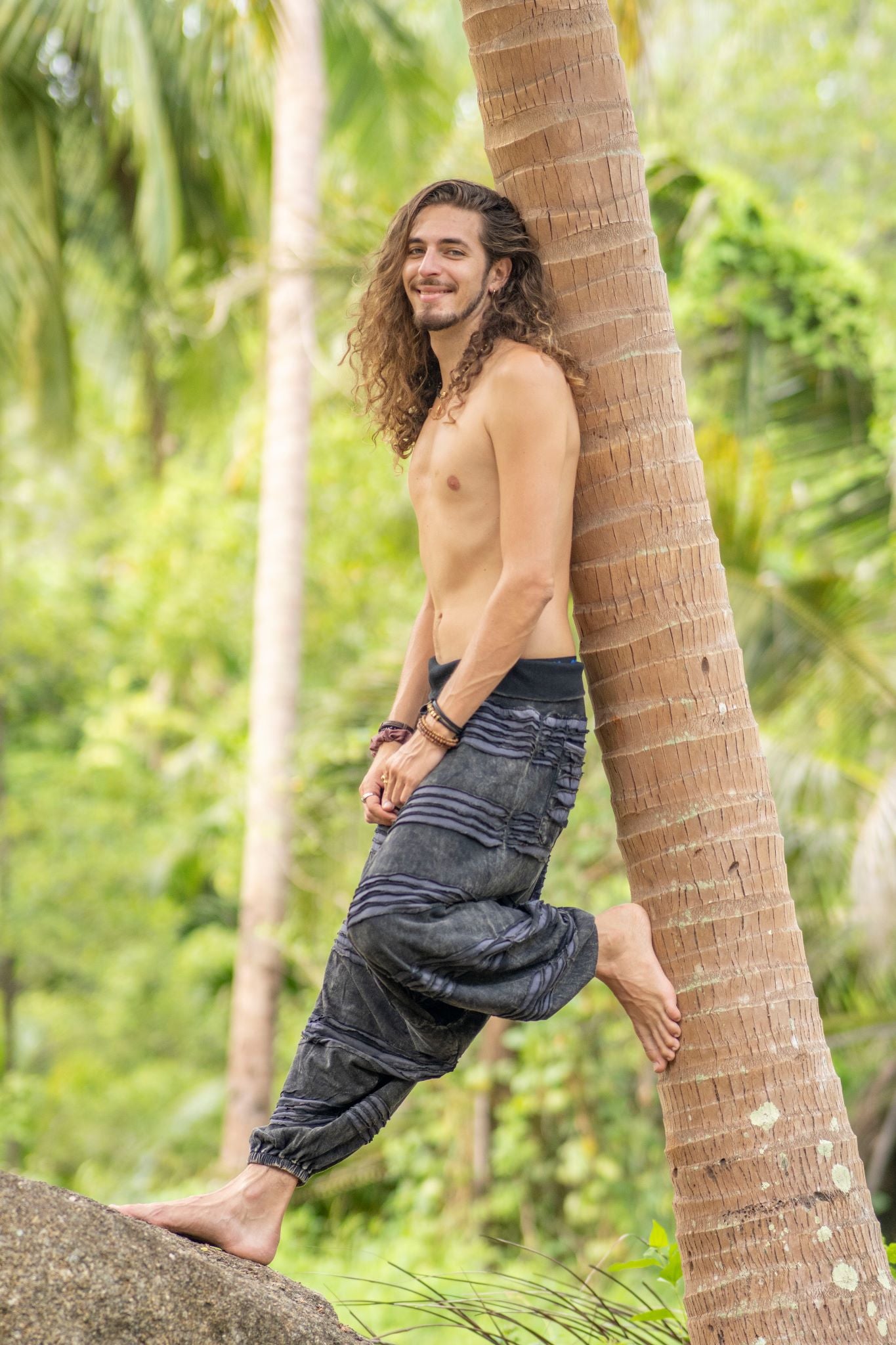 Man wearing grey stonewashed cotton razorcut hippie trousers with a relaxed drop crotch fit, leaning against a palm tree in a tropical bohemian setting.