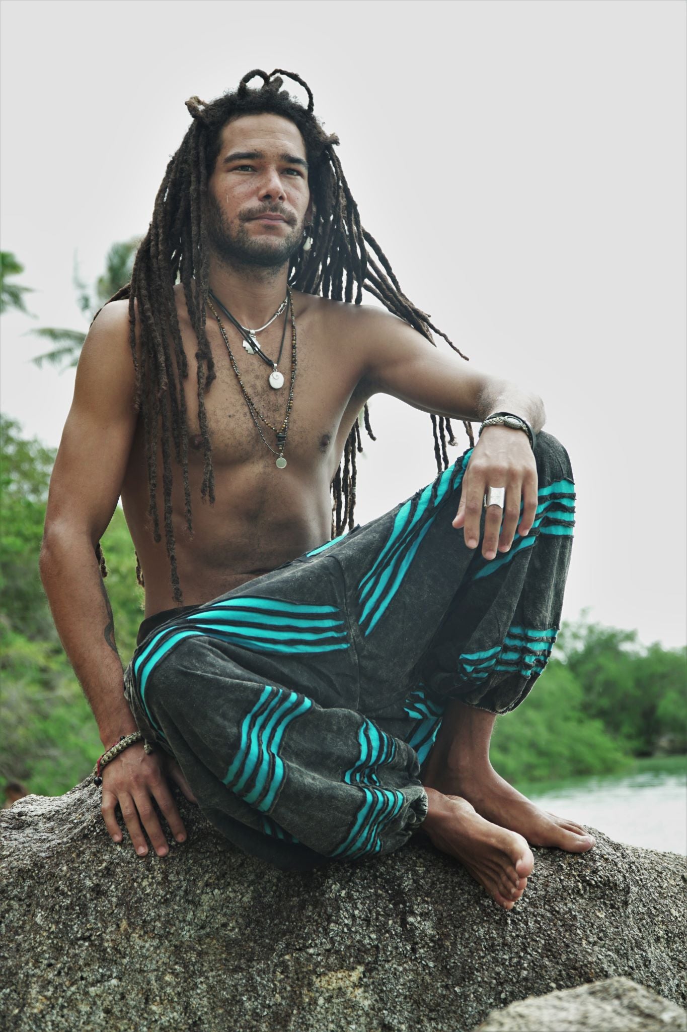 Male model with long dreadlocks sitting cross-legged on a rock wearing charcoal stonewashed bohemian trousers with a bright blue razorcut underlayer, in a relaxed lifestyle pose with a tropical backdrop.