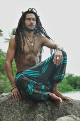 Male model with long dreadlocks sitting cross-legged on a rock wearing charcoal stonewashed bohemian trousers with a bright blue razorcut underlayer, in a relaxed lifestyle pose with a tropical backdrop.