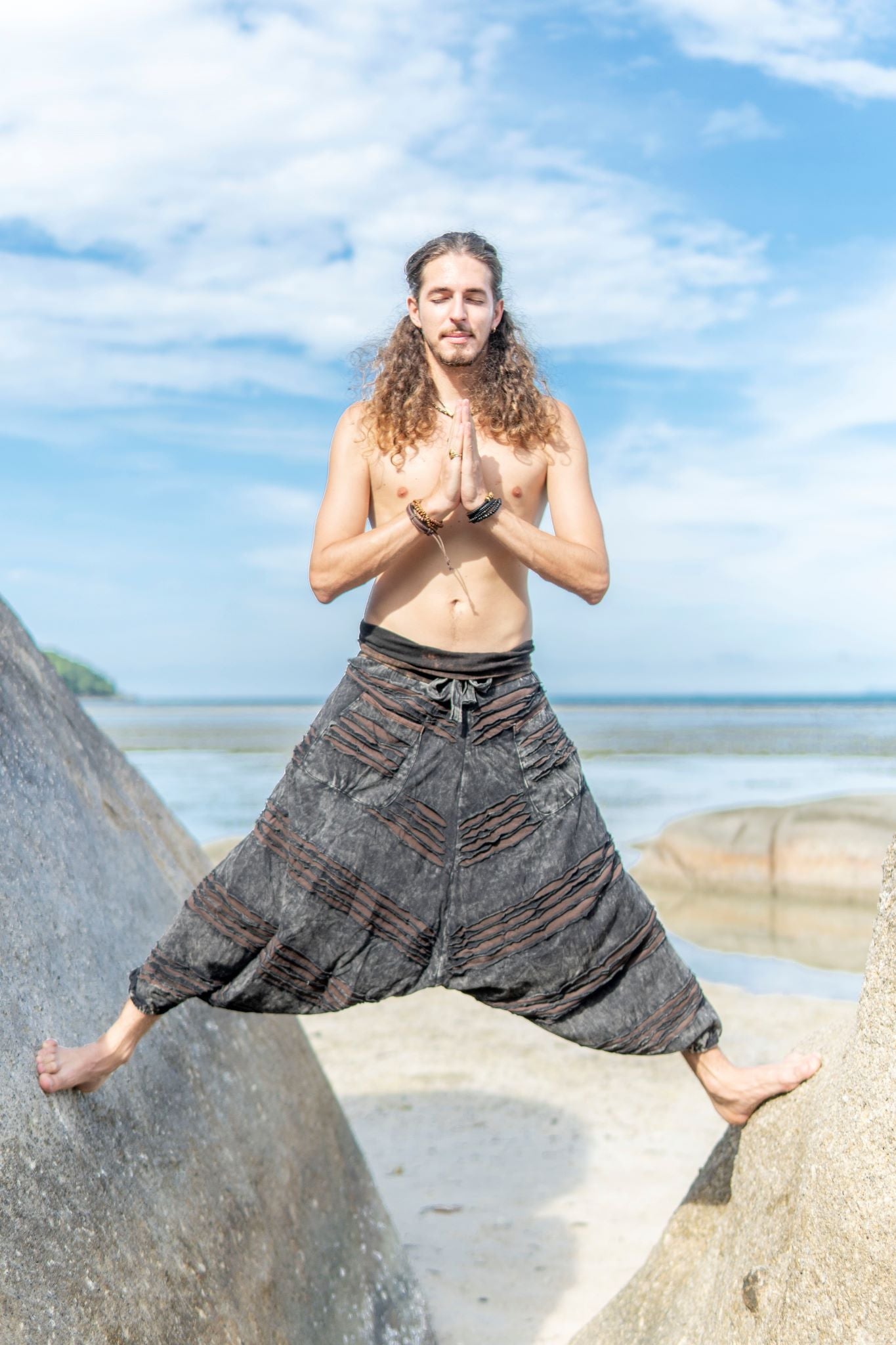 Man wearing charcoal grey stonewashed men's harem pants with a brown razorcut underlay in a yoga-inspired pose on tropical beach rocks.