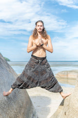 Man wearing charcoal grey stonewashed men's harem pants with a brown razorcut underlay in a yoga-inspired pose on tropical beach rocks.