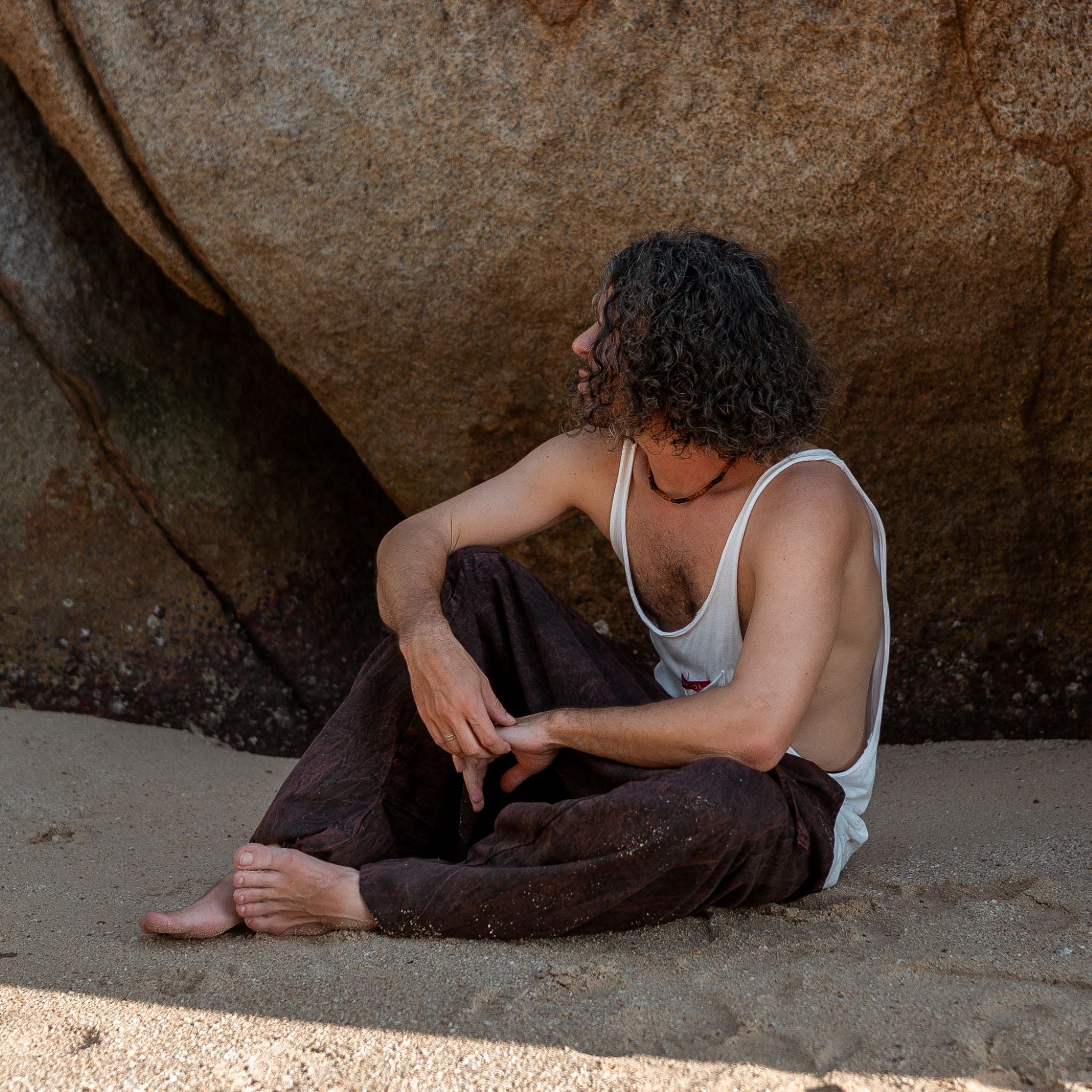 Man sitting on sand wearing brown stonewashed drop crotch harem pants, boho festival lifestyle