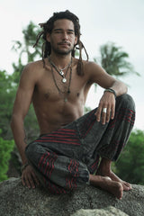 Man sitting on a rock wearing charcoal grey stonewashed hippie harem pants with maroon razorcut detailing in a tropical setting.