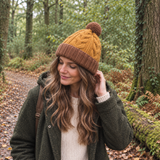 Woman wearing a chunky knit wool hat in mustard yellow/orange with a brown upturned rim and matching pom pom. The hat is lined with fleece and has a cableknit design