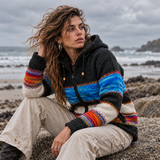 woman sitting on a beach rock wearing a colourful fleece lined wool coat with a blue tiee panel and a rainbow one.